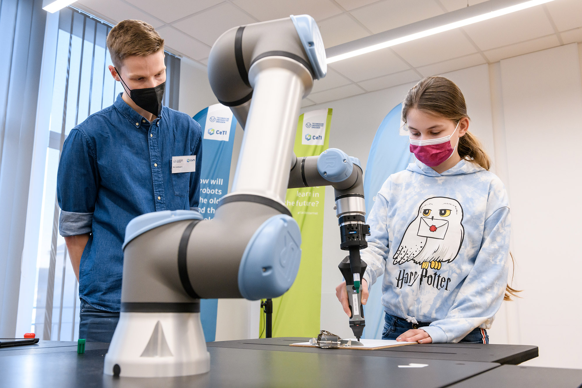Photograph of a child next to a robotic arm while using a smart pen. Next to the child is a researcher explaining the functionality of the robot.