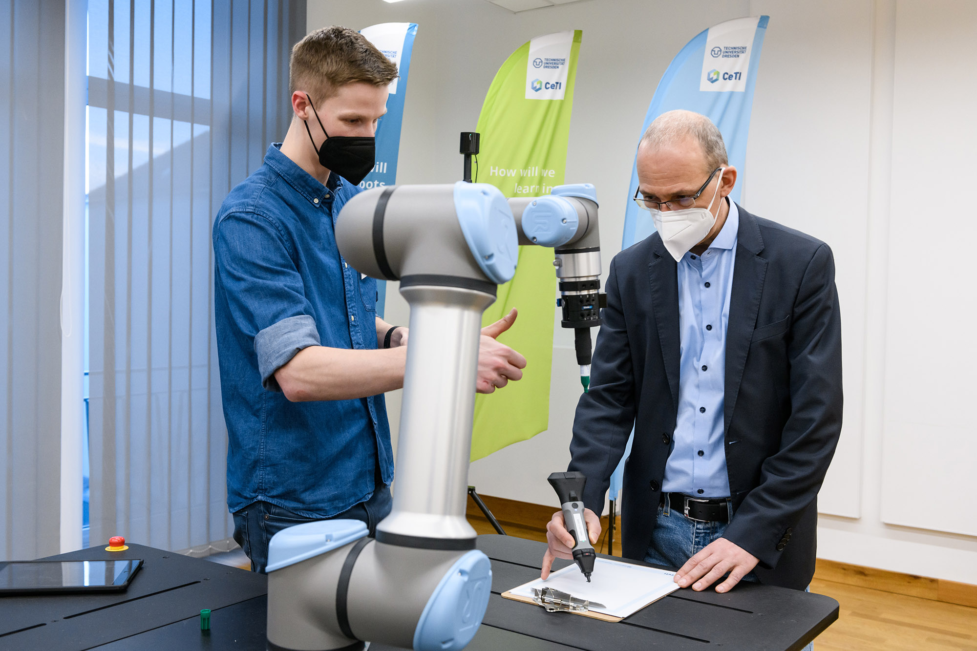 Photograph of two men next to a robotic arm, one of them is holding a smart pen