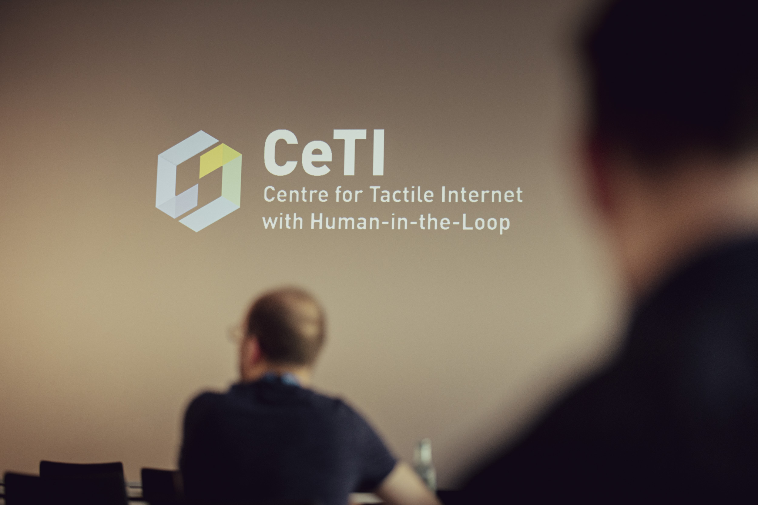 Photograph of projection onto a wall with writing''CeTI Centre for Tactile Internet with human-in-the-Loop" with two people visible from behind