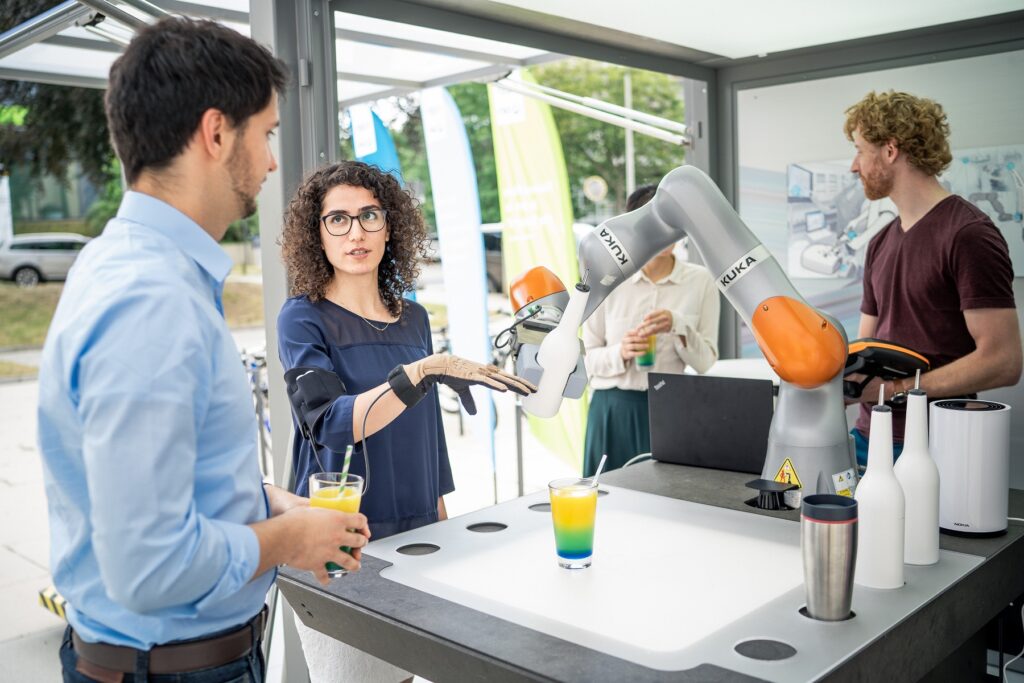 A woman standing next to a robotic arm while wearing a connected glove and explaining the functionality of the robot to a man next to her.