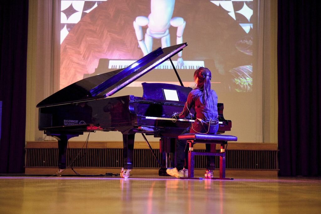 A person playing a grand piano while being connected to a device. Behind the piano is a projection of the digital avatar mirroring the person playing the piano.