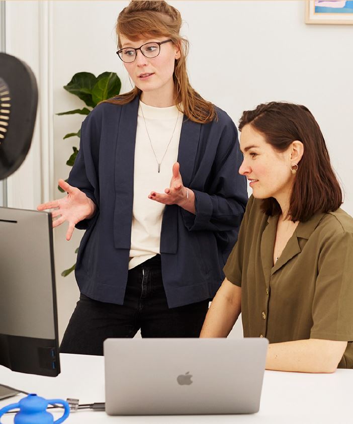 image of two women. One is sitting at a desk and the other is talking to him while looking at a computer