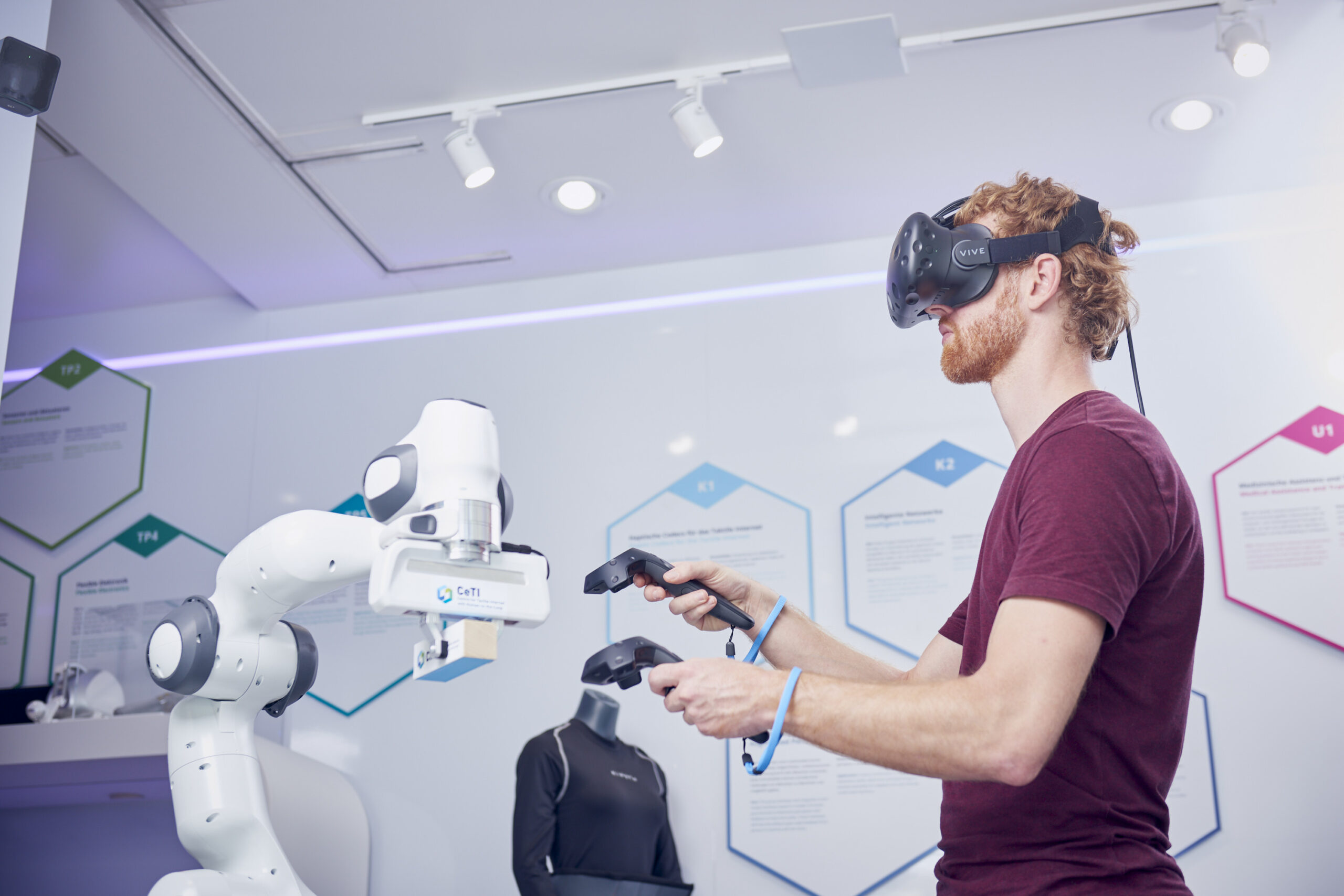 Ein Mann mit einer Virtual-Reality-Brille und zwei Controllern in der Hand. Neben dem Mann sehen wir einen Roboterarm.