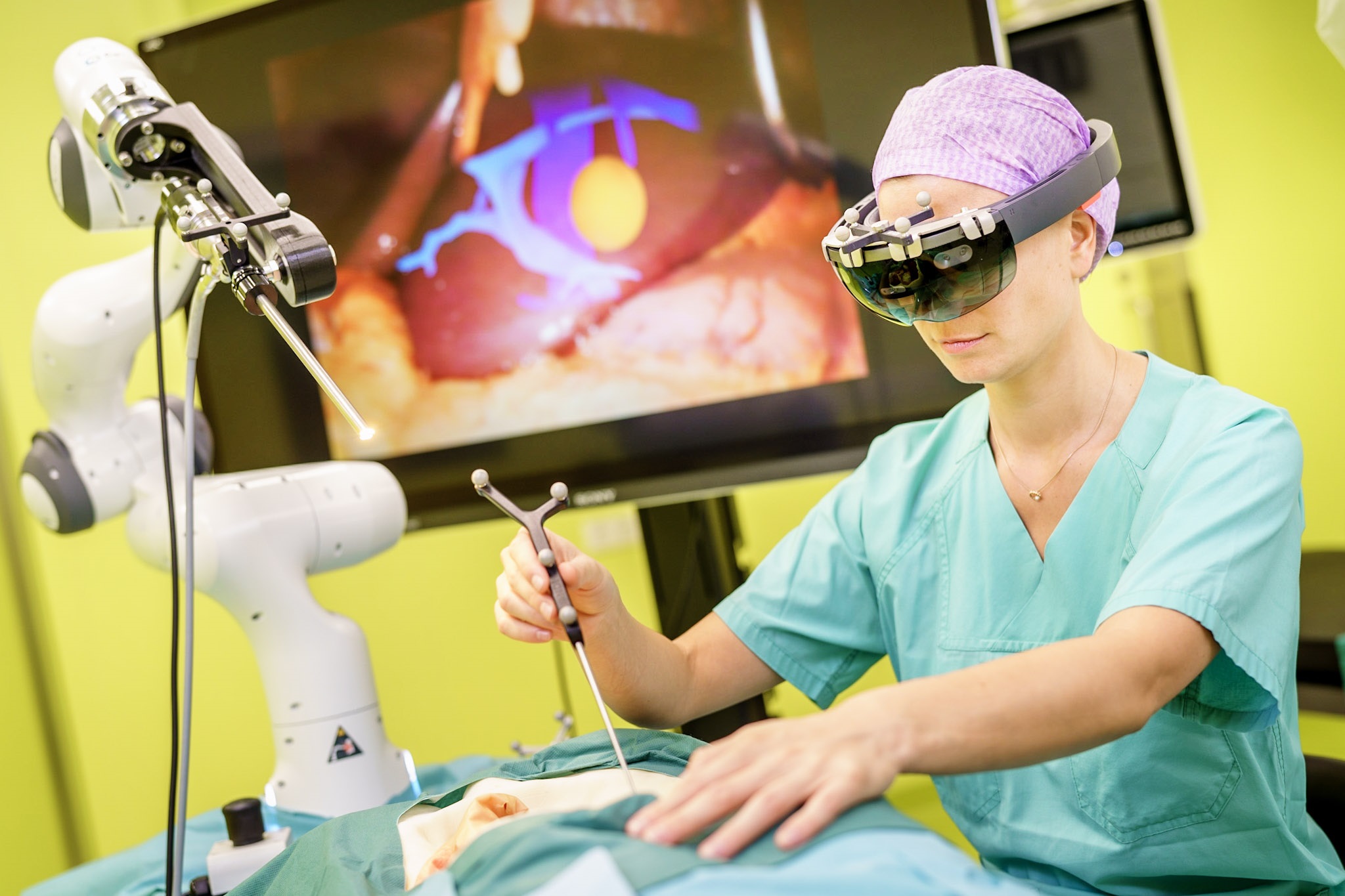 Ein Arzt operiert einen Plastikkörper und wird dabei von einem Roboterarm unterstützt. Hinter dem Arzt ist ein Bildschirm mit menschlichen Organen zu sehen.