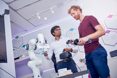 Zwei Forscher benutzen einen Roboterarm. Der eine hält Controller in der Hand, der andere einen Virtual-Reality-Visor.