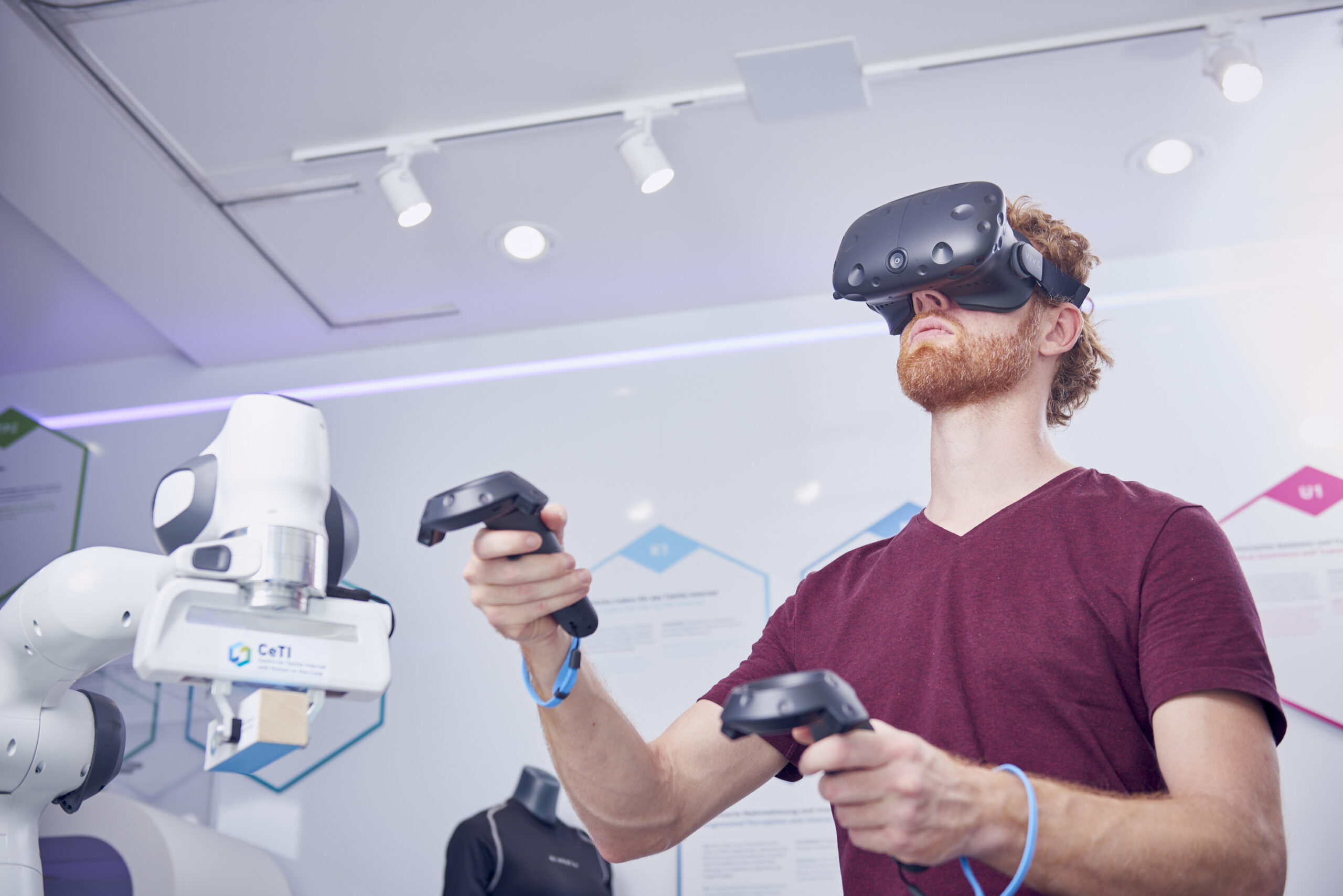 A man wearing a virtual reality visor and holding two controllers. Next to the man we see a robotic arm.