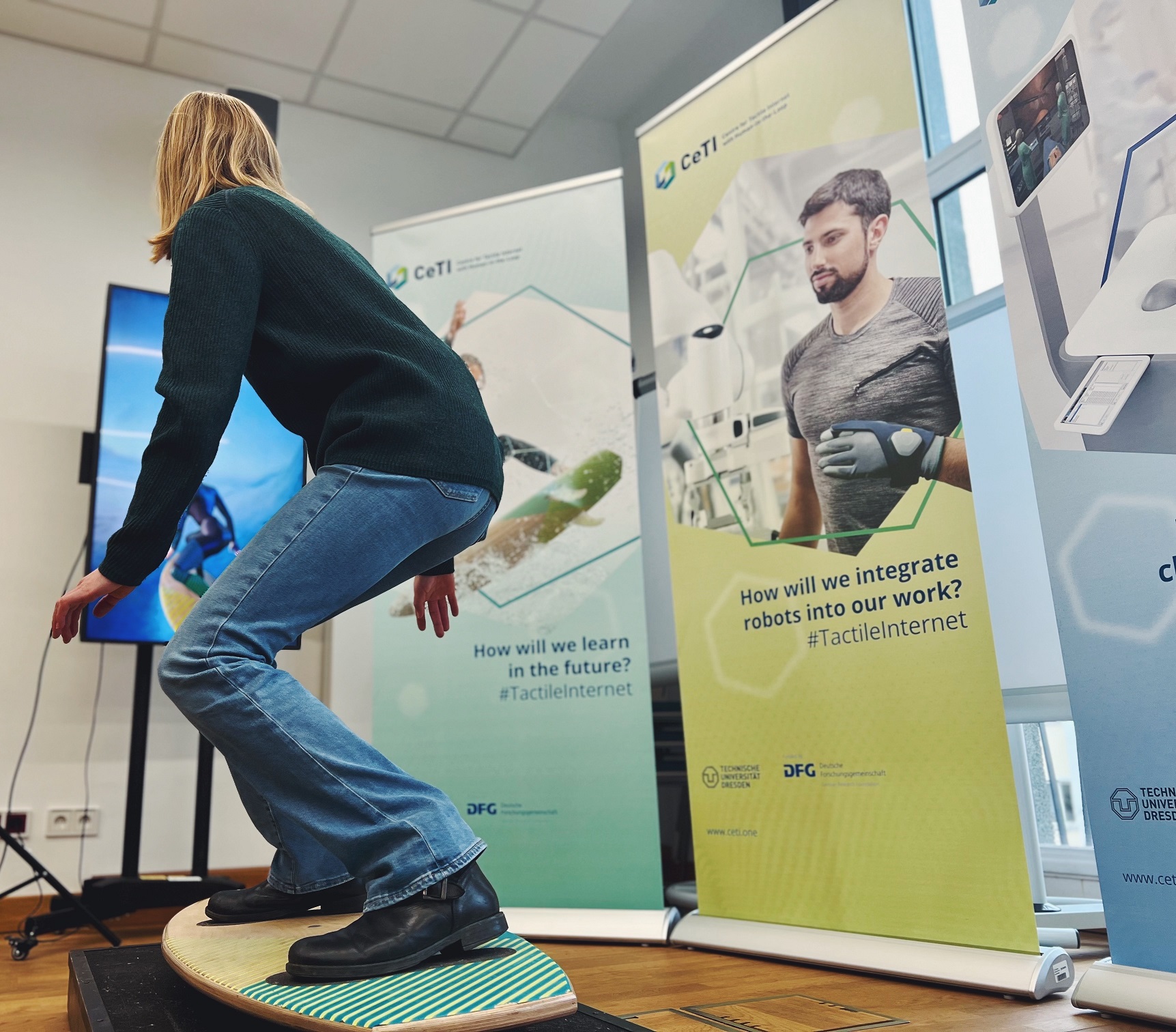 Photograph of a person in a room standing on a surfboard with a screen in front of them with a digital avatar copying the movements